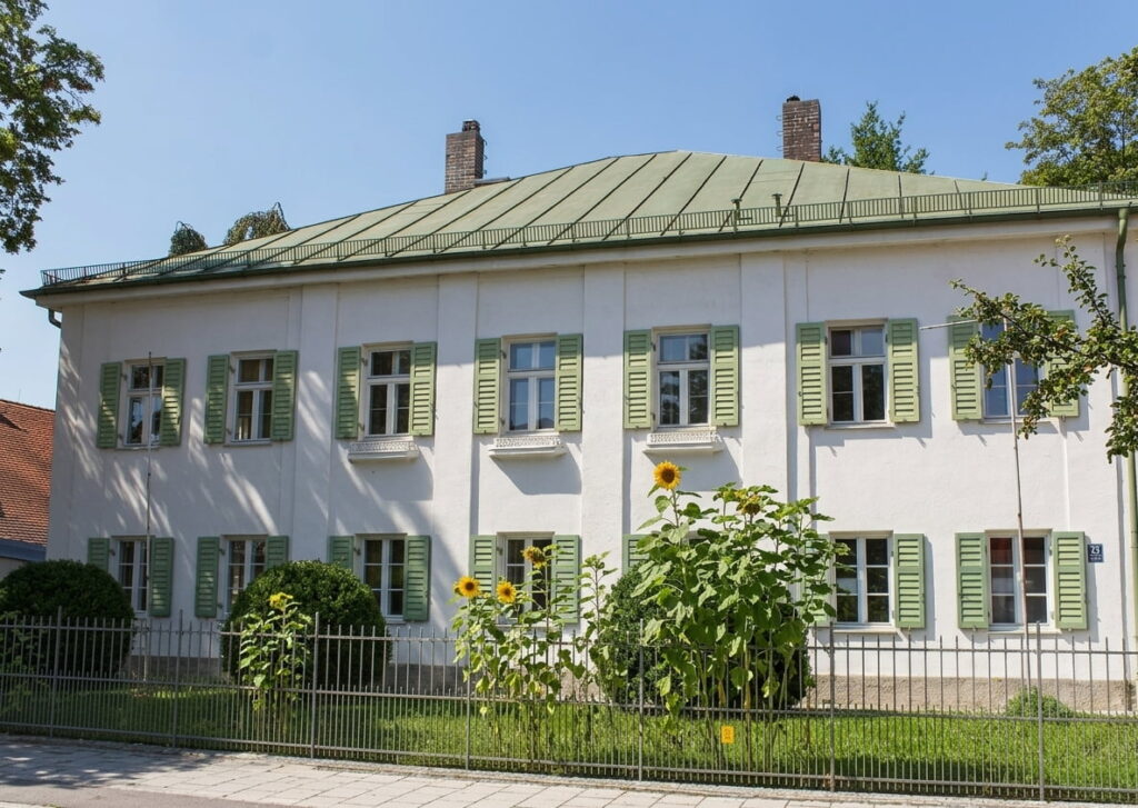 Zweigeschossiges weißes Wohnhaus mit grün lackierten Fensterläden und grünem Blechdach hinter einem niedrigen Metallzaun mit Sonnenblumen davor