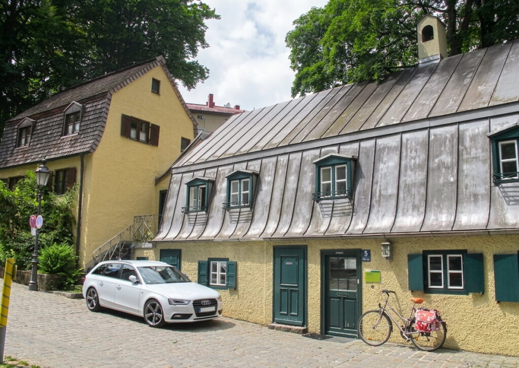 Zweistöckiges gelbes Wohnhaus mit grünem Tür- und Fensterrahmen, davor ein weißer Audi-Kombi und ein Fahrrad mit rotem Korb an einer Kopfsteinpflasterstraße in Haidhausen.