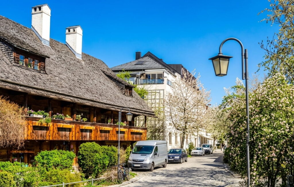 Straße in Haidhausen mit traditionellem Fachwerkhaus links, mehreren parkenden Autos und blühenden Bäumen rechts