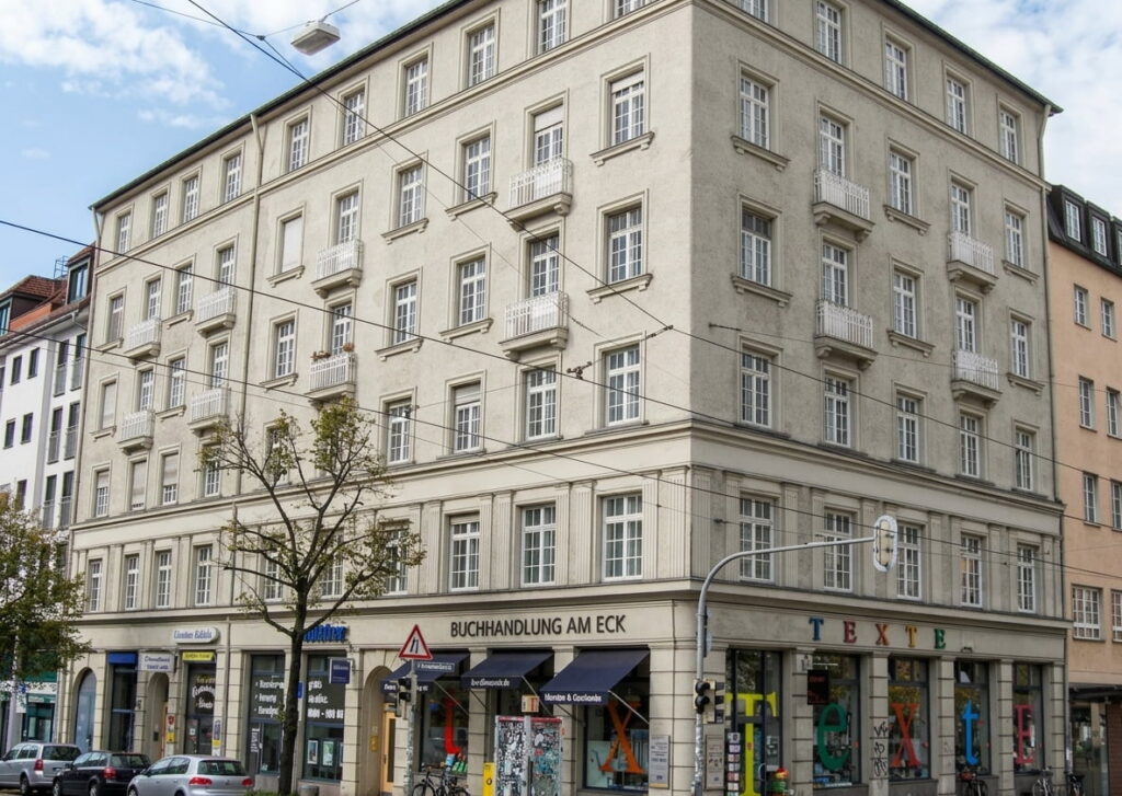 Mehrstöckiges Eckgebäude mit klassischer Fassade und mehreren Ladengeschäften im Erdgeschoss, darunter eine Buchhandlung mit dem Schriftzug 'BUCHHANDLUNG AM ECK'