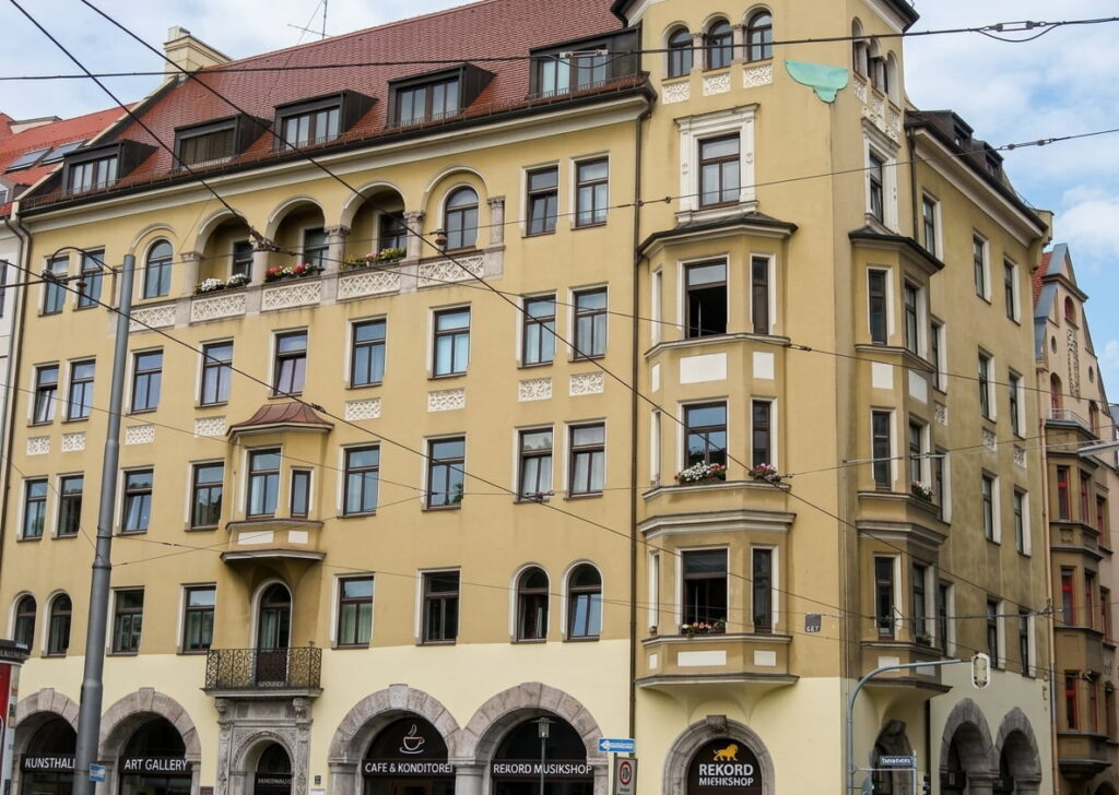 Mehrstöckiges Eckgebäude mit gelber Fassade, Arkaden im Erdgeschoss und mehreren Fenstern, darunter ein Erker an der Ecke