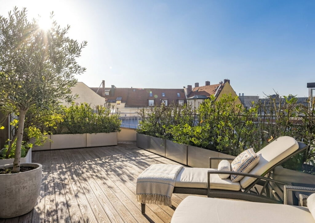 Dachterrasse mit Holzdeck, Liegestuhl mit Kissen und Decke, großen Pflanzkübeln mit Sträuchern und Baum, im Hintergrund Häuserdächer unter klarem Himmel