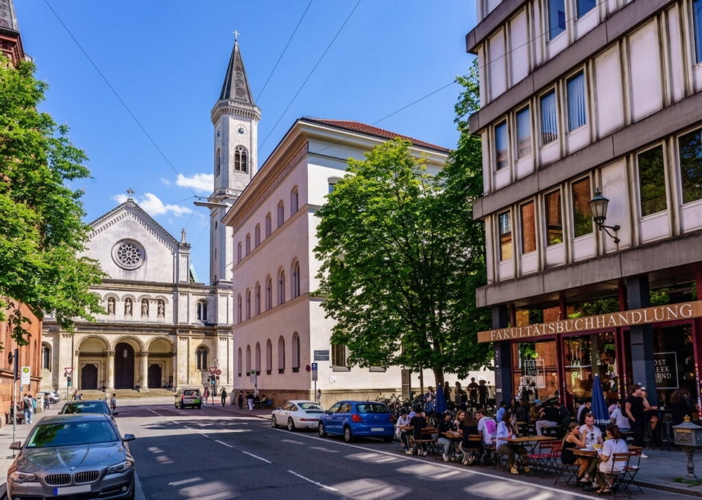 Straße in Maxvorstadt mit der Kirche St. Ludwig im Hintergrund und einer belebten Außengastronomie neben einem Gebäude mit dem Schild 'Fakultätsbuchhandlung'