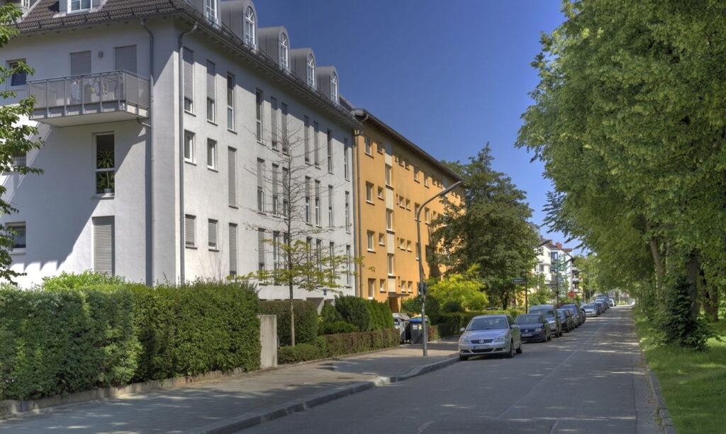 Straße in einem Wohngebiet mit mehrstöckigen Wohnhäusern links und parkenden Autos rechts unter blauem Himmel