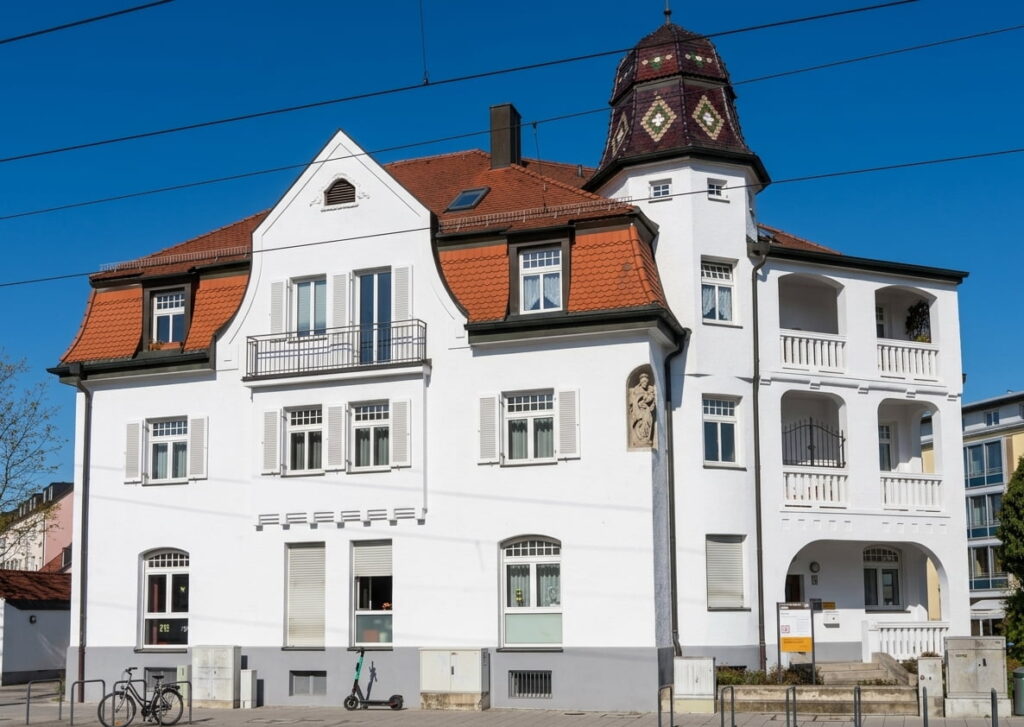 Weißes mehrstöckiges Wohngebäude mit rotem Satteldach, Erker mit Balkon und rundem Turm mit Ziegeldach vor blauem Himmel