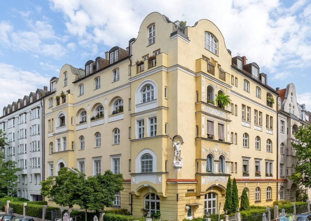 Mehrstöckiges Eckgebäude mit gelber Fassade, Erkern, Rundbogenfenstern und einer Statue an der Ecke, umgeben von Bäumen und angrenzenden Wohnhäusern