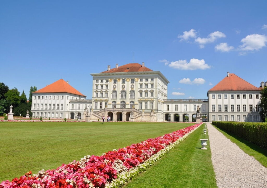 Barockschloss mit drei Gebäudeteilen, davor ein breiter Rasen mit einem Blumenbeet und einem Kiesweg, blauer Himmel mit wenigen Wolken