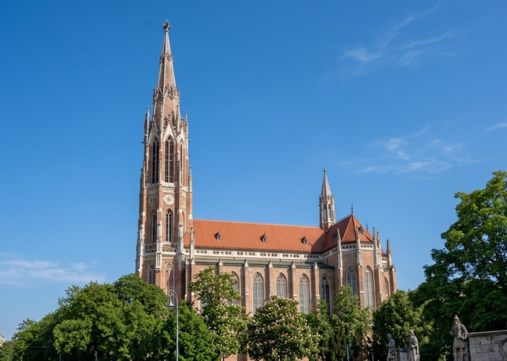 Gotische Kirche mit hohem Turm und roten Dachziegeln vor blauem Himmel und Bäumen im Vordergrund