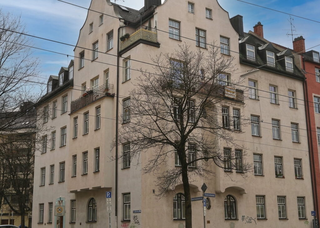 Mehrstöckiges Wohngebäude mit Satteldach und mehreren Fenstern an einer Straßenecke, davor ein kahler Baum und Verkehrsschilder