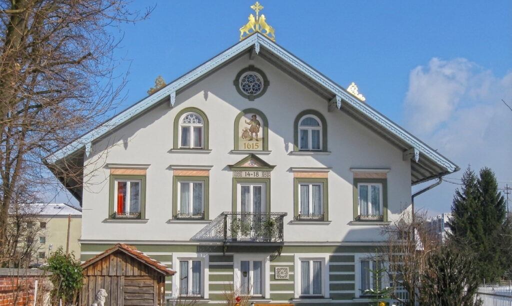 Fachwerkhaus mit Giebel, Balkonen und historischen Verzierungen, umgeben von Bäumen und einem kleinen Holzschuppen im Vordergrund