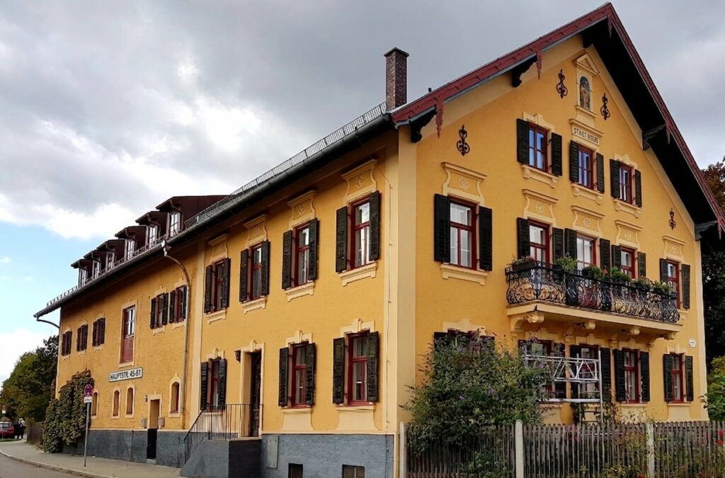 Mehrstöckiges gelb gestrichenes Wohnhaus mit roten Fensterläden und einem Balkon mit Blumenkästen an einer Straßenecke unter bewölktem Himmel