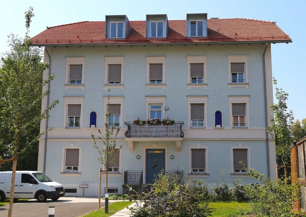 Dreigeschossiges Wohnhaus mit rotem Satteldach, mehreren Fenstern und einem Balkon mit Blumenkästen, davor ein weißer Transporter und kleine Bäume auf einer Rasenfläche