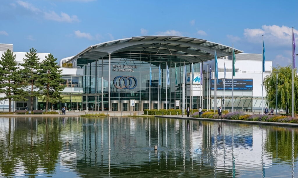 Großes modernes Messegebäude mit Glasfassade und geschwungenem Dach vor einem Teich mit Spiegelung