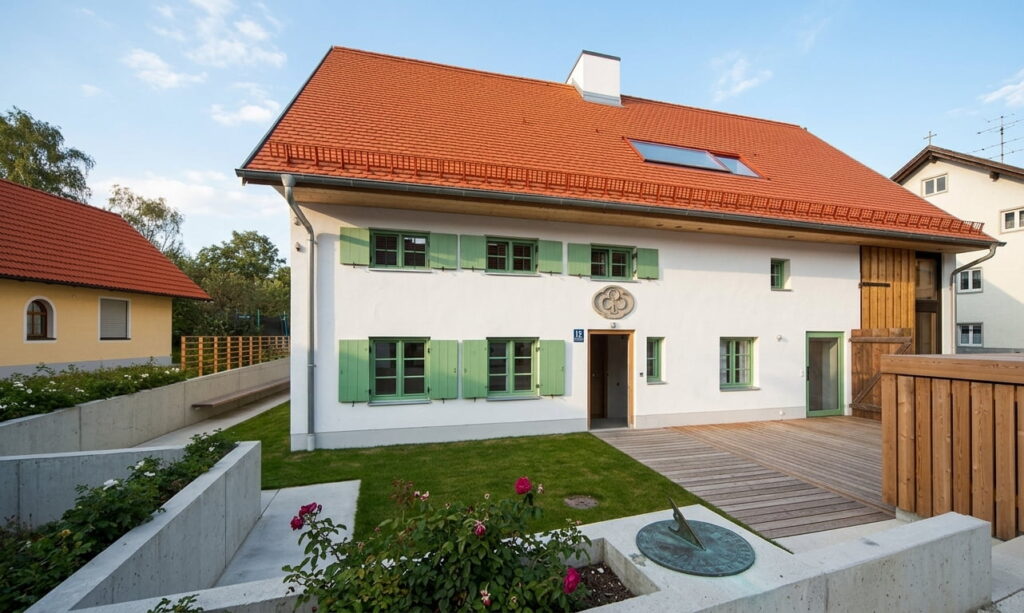 Zweistöckiges weißes Haus mit grün lackierten Fensterläden, rotem Satteldach und Holzveranda vor grünem Garten mit Rosen und Sonnenuhr