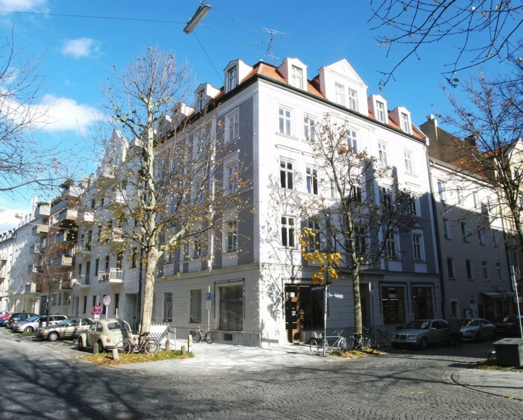 Eckgebäude mit mehreren Stockwerken und Mansardendach an einer ruhigen Straße mit geparkten Autos und kahlen Bäumen im Stadtviertel Schwabing-West