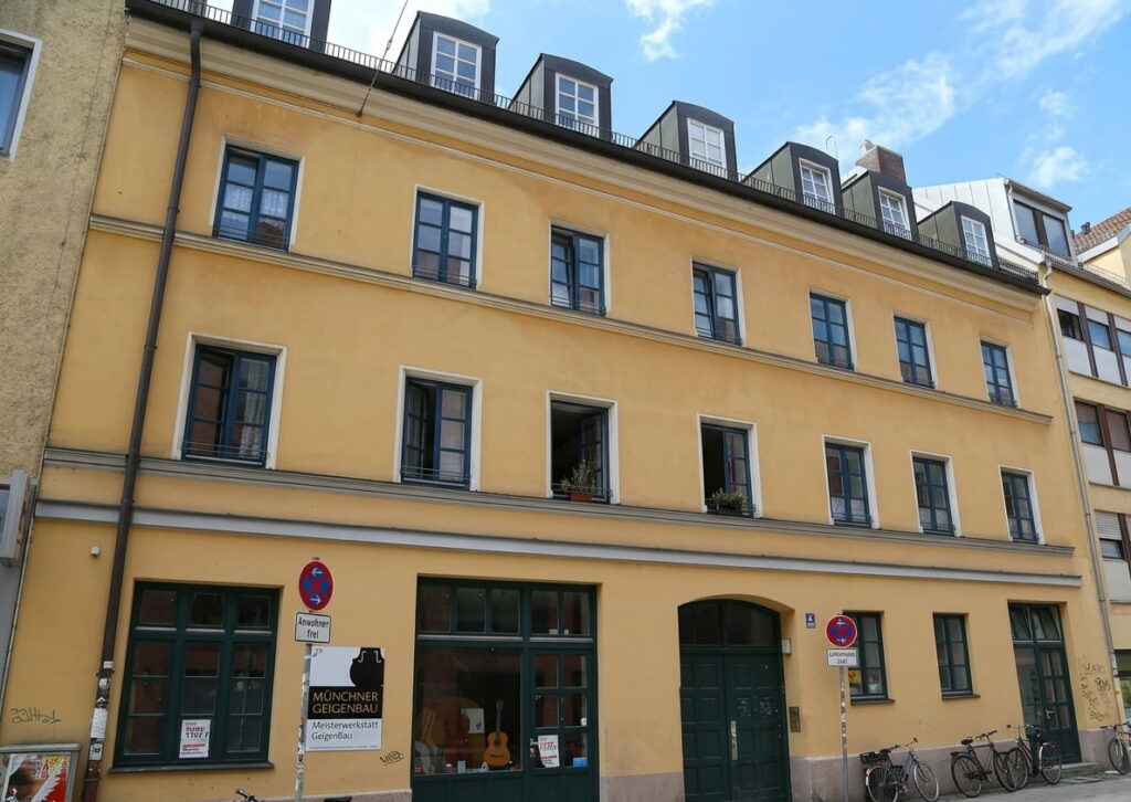 Mehrstöckiges gelbes Wohngebäude mit mehreren Fenstern, zwei großen grünen Türen und einem Schild mit der Aufschrift 'Münchner Gegenwart' an der Fassade