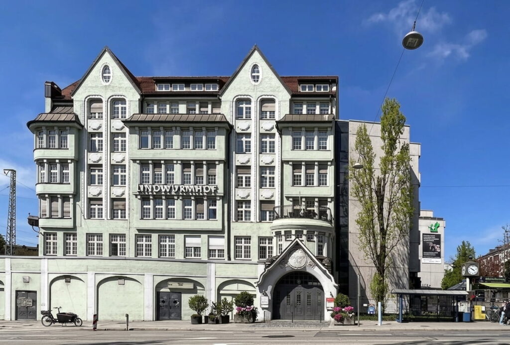Mehrstöckiges historisches Gebäude mit dem Schriftzug 'INDUSTRIEHOF' an der Fassade, davor Straße mit parkenden Motorrädern und Bäumen, blauer Himmel im Hintergrund