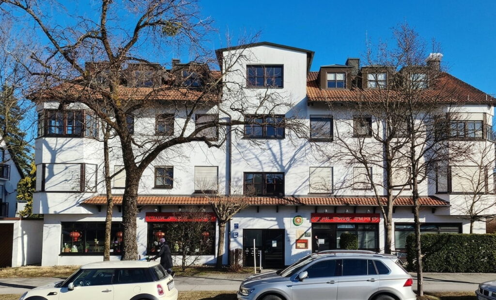 Mehrstöckiges Wohn- und Geschäftshaus mit weißen Fassaden, roten Dachziegeln, mehreren Fenstern und zwei geparkten Autos davor