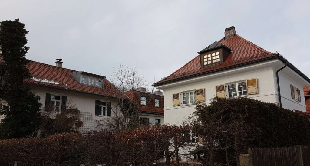 Zwei Wohnhäuser mit Satteldächern und Dachgauben hinter kahlen Büschen unter bewölktem Himmel in Trudering