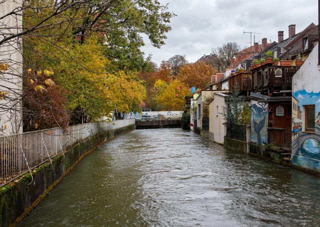 Blick auf einen Fluss mit herbstlich gefärbten Bäumen links und Wohnhäusern mit Balkonen rechts