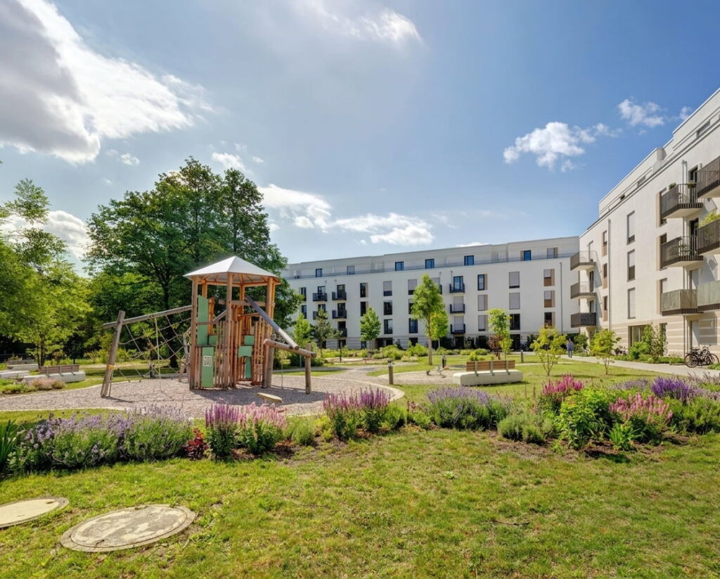 Moderner Wohnkomplex mit mehrstöckigen Gebäuden, zentralem Spielplatz und gepflegtem Grünbereich unter blauem Himmel