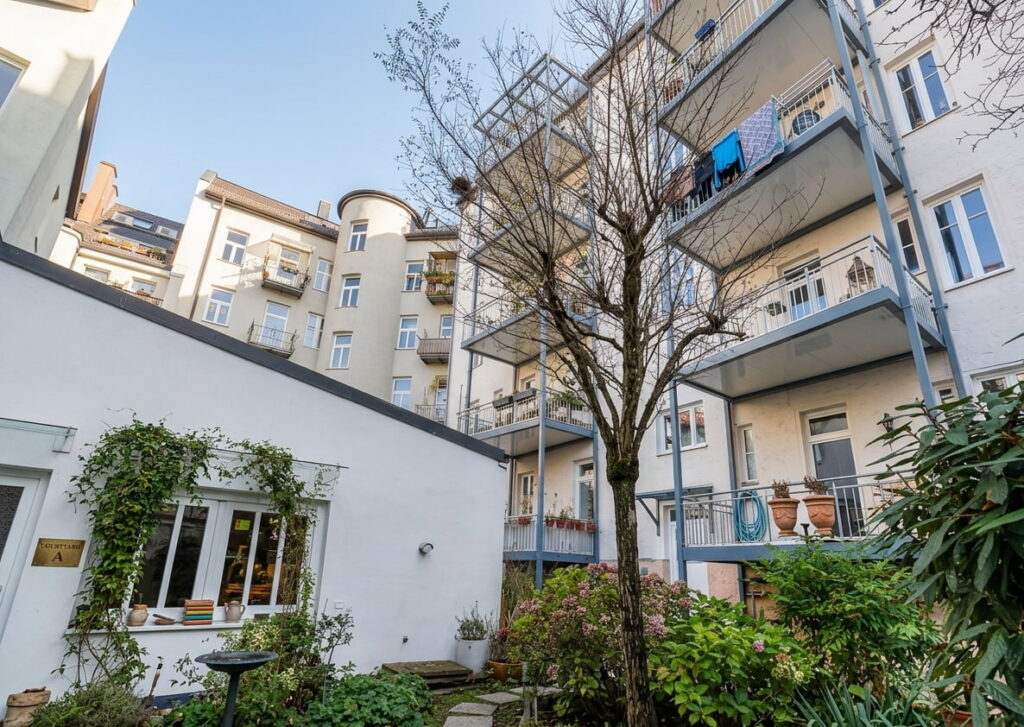 Innenhof mit kleinem Garten, umgeben von mehrstöckigen Wohngebäuden mit Balkonen und einem Baum in der Bildmitte