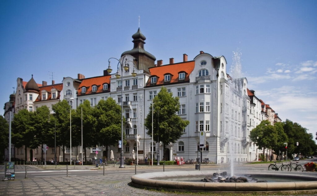 Historisches Eckgebäude mit roten Dächern und Türmchen in Bogenhausen vor blauem Himmel mit Springbrunnen und Bäumen im Vordergrund