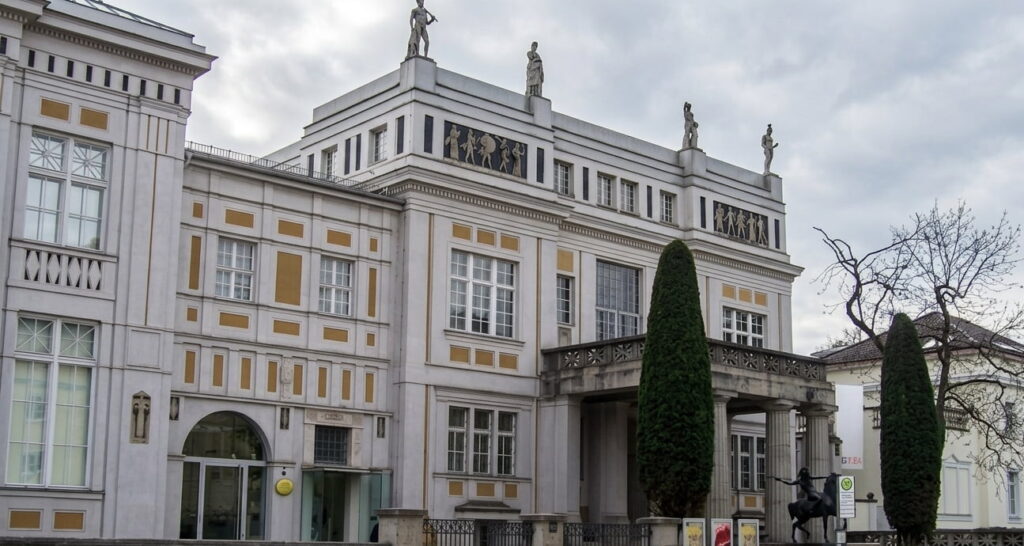 Fassade eines historischen Gebäudes mit Säulen, Statuen auf dem Dach und zwei hohen, schmalen Bäumen davor