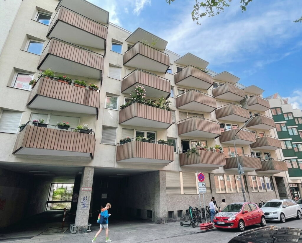 Mehrstöckiges Wohngebäude mit mehreren Balkonen und Blumenkästen in einem städtischen Viertel, darunter ein Durchgang und geparkte Autos auf der Straße
