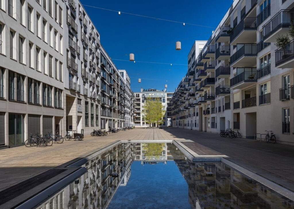 Blick auf einen modernen Innenhof mit Wasserbecken, umgeben von mehrstöckigen Wohngebäuden mit Balkonen und Fahrradständern unter blauem Himmel