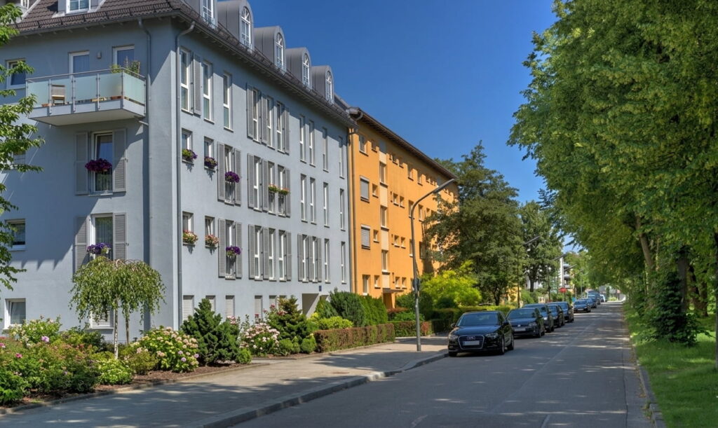 Mehrstöckige Wohnhäuser in Milbertshofen mit Balkonen und parkenden Autos an einer von Bäumen gesäumten Straße