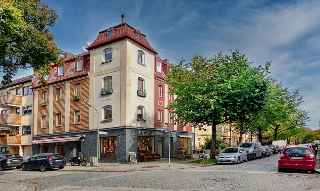 Eckgebäude mit Café 'THE DAILY GRIND' im Erdgeschoss an einer von Bäumen gesäumten Straße mit geparkten Autos in Milbertshofen
