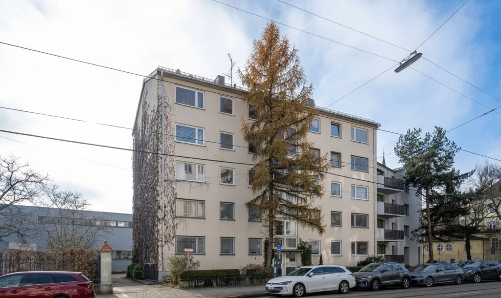 Mehrstöckiges Wohnhaus mit mehreren Fenstern und Bäumen davor, geparkte Autos am Straßenrand