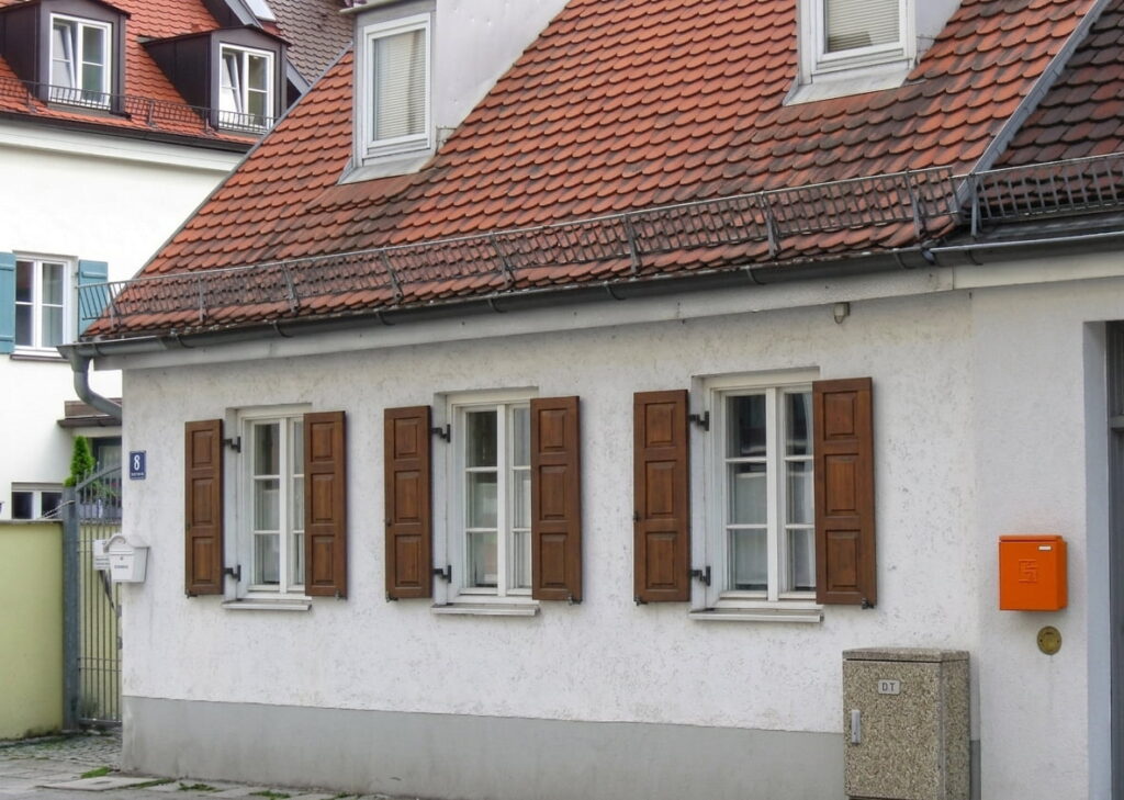 Einfamilienhaus mit weißer Fassade, braunen Fensterläden und rotem Ziegeldach in einer Wohnstraße