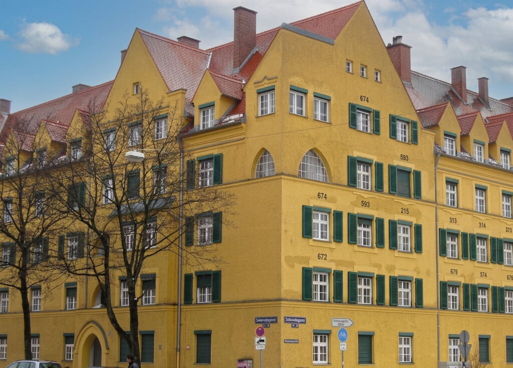 Mehrstöckiges gelbes Wohngebäude mit grünen Fensterläden und roten Satteldächern in Ramersdorf