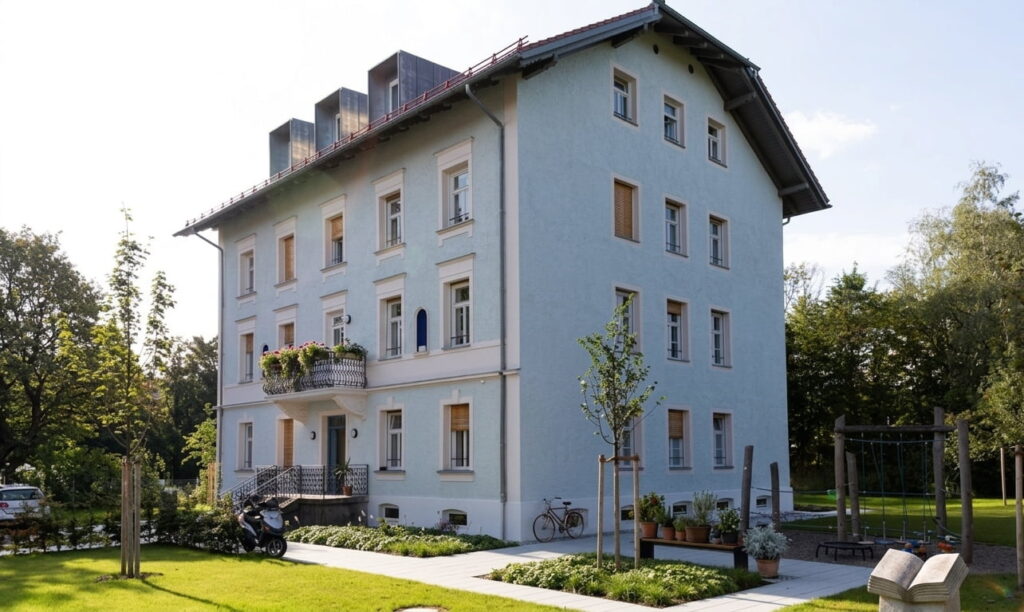 Mehrstöckiges Wohnhaus mit weißer Fassade, Balkon mit Blumen und umgeben von gepflegtem Garten in Ramersdorf
