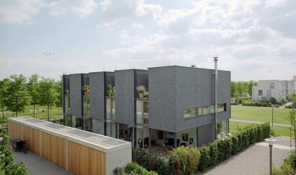 Modernes, kubisches Wohngebäude mit dunkler Fassade und großen Fenstern in einer grünen Wohnanlage in Riem