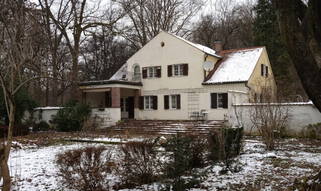 Zweistöckiges Haus mit Satteldach, mehreren Fenstern mit Klappläden und einer Terrasse, umgeben von kahlen Bäumen und leicht verschneitem Garten in Schwabing-West