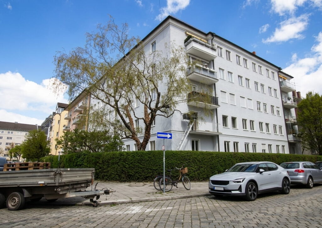 Mehrstöckiges Wohnhaus mit Balkonen an der Straßenecke, davor geparkte Autos und ein Fahrrad, im Vordergrund ein Baum und eine Hecke