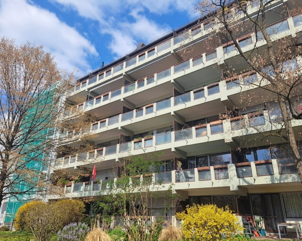 Mehrstöckiges Wohngebäude mit Balkonen und Glasgeländern vor blauem Himmel und Bäumen mit herbstlichem Laub
