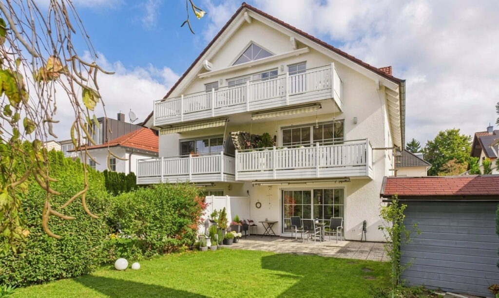 Zweistöckiges weißes Wohnhaus mit Balkon und Terrasse, umgeben von grünem Garten und Hecke, bei sonnigem Himmel