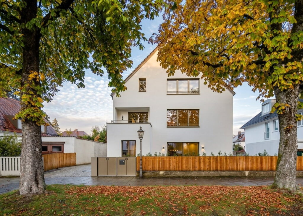 Modernes dreistöckiges Wohnhaus mit weißer Fassade, großen Fenstern und Holzzaun, umgeben von herbstlich gefärbten Bäumen