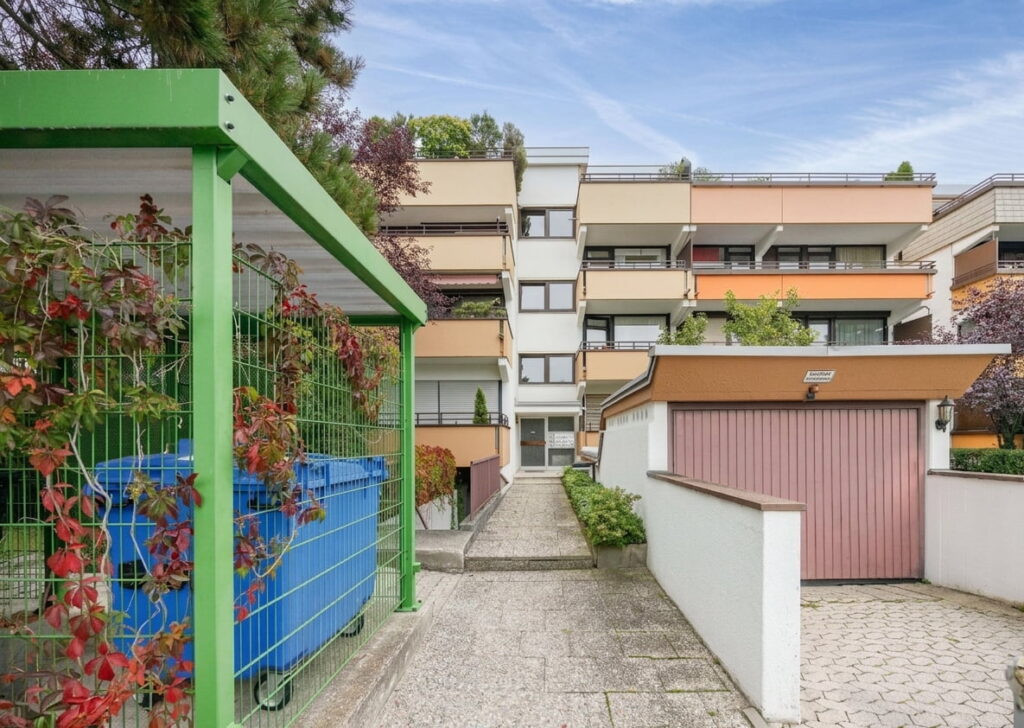 Mehrstöckiges Wohngebäude mit Balkonen und einem kleinen Vorgartenbereich neben einem Weg, links ein grüner Metallzaun mit Pflanzen und blauen Müllcontainern, rechts eine Garage mit brauner Tür.
