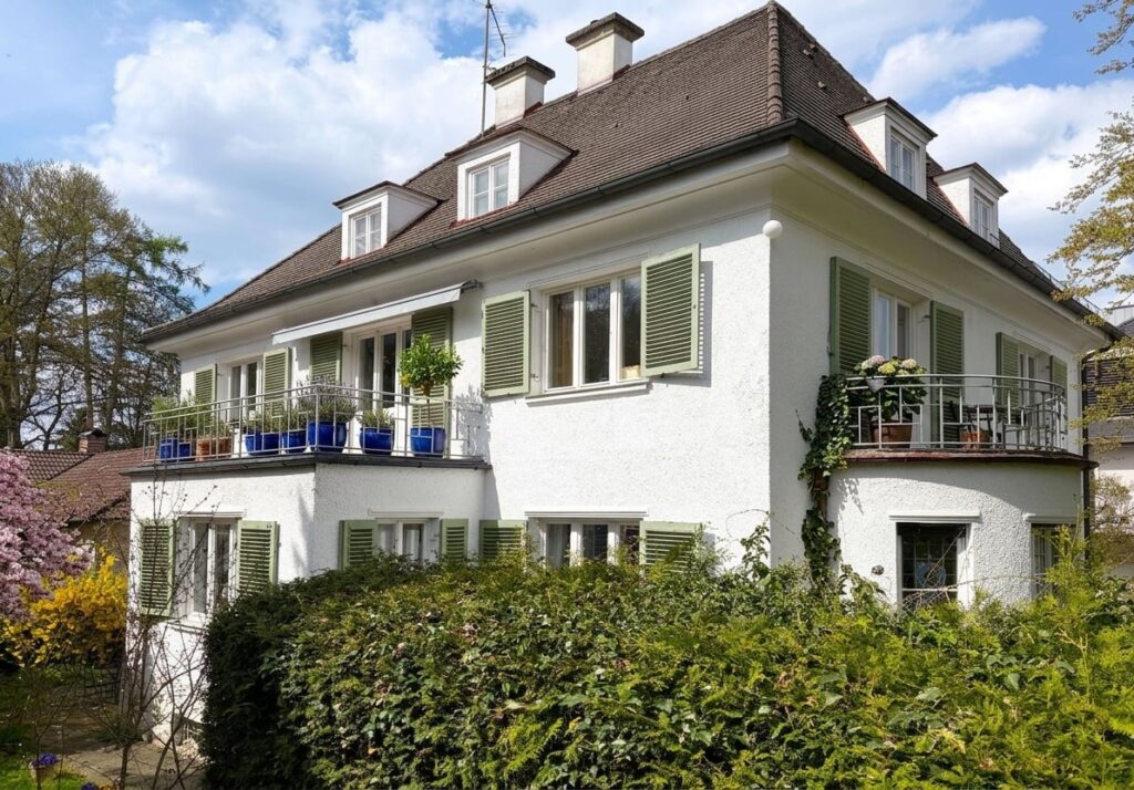 Großes weißes Wohnhaus mit grün lackierten Fensterläden und Balkon in einem grünen Garten