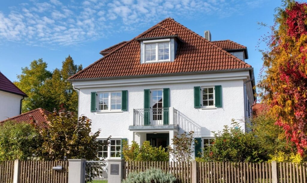 Zweistöckiges weißes Haus mit rotem Ziegeldach, grünen Fensterläden und Holzzaun im Garten in Harlaching