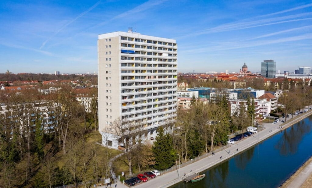 Hochhaus mit Balkonen an einem Flussufer in Thalkirchen, umgeben von Bäumen und weiteren Gebäuden im Hintergrund
