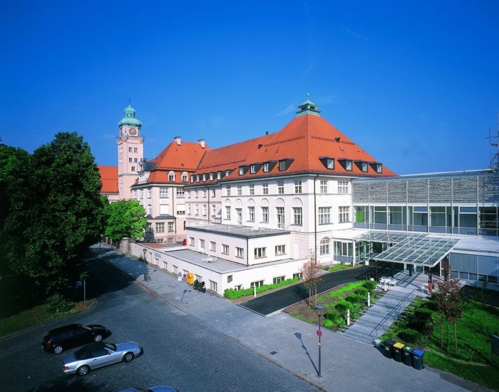 Großes historisches Gebäude mit rotem Walmdach und Uhrturm bei klarem Himmel in Harlaching