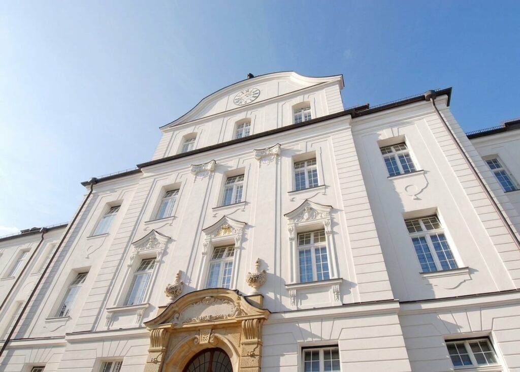 Fassade eines mehrstöckigen historischen Gebäudes mit weißen Wänden, verzierten Fensterrahmen und einem kunstvoll gestalteten Eingang unter blauem Himmel
