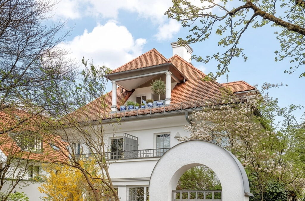 Mehrstöckiges Wohnhaus mit Satteldach, Balkon mit Pflanzen und weißem Torbogen im Vordergrund, umgeben von Bäumen und Sträuchern