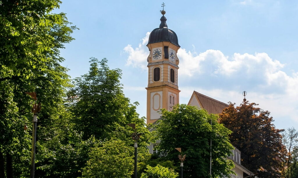 Kirchturm mit Ziffernblatt und Zwiebeldach hinter grünen Bäumen bei blauem Himmel in Thalkirchen